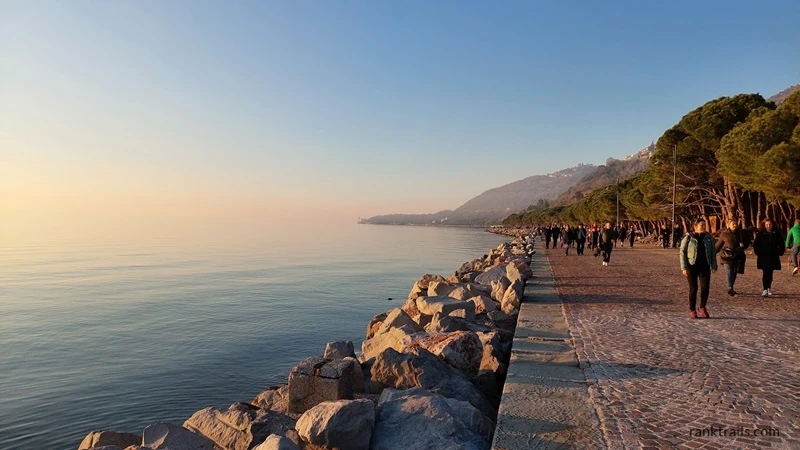 People walking along Barcola promenade in Trieste at sunset with sea views and rocky coastline.