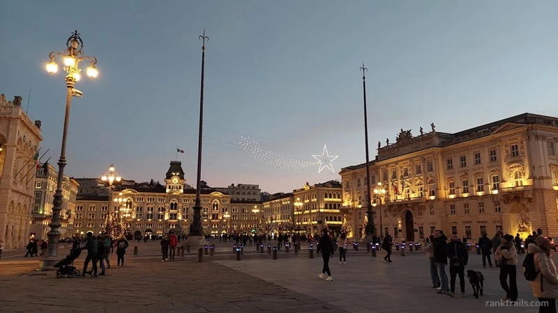 Evening view of Piazza Unità d’Italia in Trieste with illuminated historic buildings and people in the square.